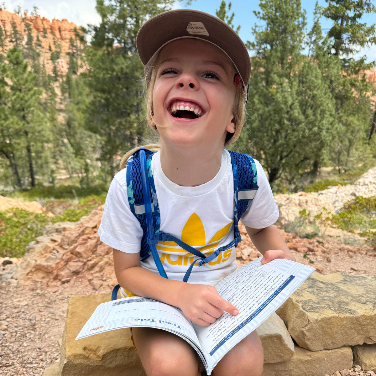 A young child sitting outdoors on a stone ledge in a natural mountain setting, holding an open book. The child is wearing a brown cap, a white T-shirt with a yellow Adidas logo, a blue backpack, and light blue hiking boots.